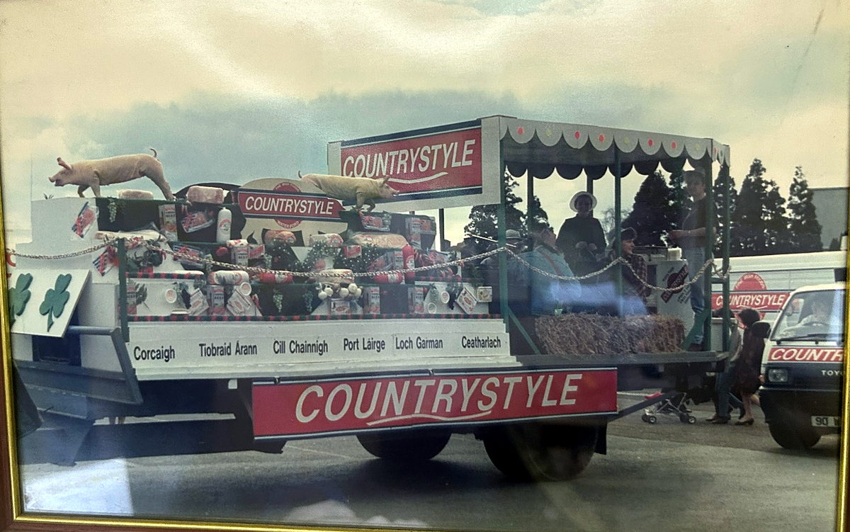 A framed photograph of the Countrystyle parade float, with the six county names in Irish along its side and a Countrystyle van behind